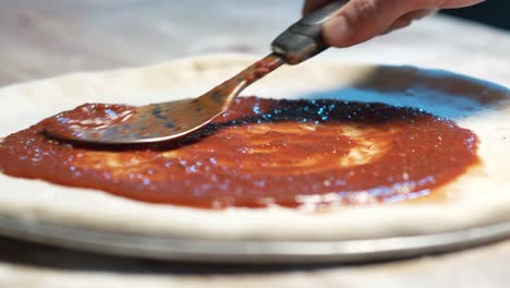 A-close-up-of-a-pizza-dough-on-a-steel-tray-on-wooden-counter-while-an-expereinced-chef-put-juicy-red-sauce-on-it-with-a-adicid