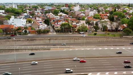 Static-drone-view-of-a-part-of-the-Panamerican-highway-where-many-cars-and-trucks-cross-it-next-to-a-neighborhood-in-Argentina-where-houses-and-tropical-trees-abound