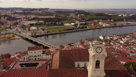 Aerial-zoom-in-University's-clock-tower-in-coimbra-cityscape-drone-view-of-the-river-and-the-bridge-with-Portugal-flag