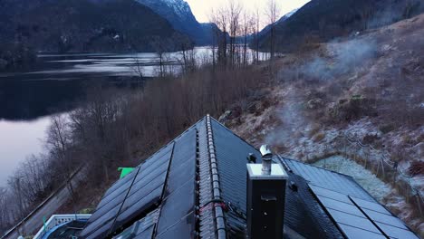 Rauch-Aus-Dem-Schornstein-Eines-Privathauses-Mit-Solarpanel-Dach-–-Geringe-Solarproduktion-–-Umgekehrte-Antenne,-Die-Das-Dach-In-Der-Nähe-Des-Schornsteins-Freigibt-–-Wunderschöne-Kalte-Fjordlandschaft-Im-Hintergrund-–-Norwegen-Antenne