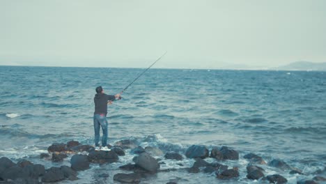 Young-man-casting-fishing-rod-standing-on-rocks-at-sea-shore-WIDE-SHOT