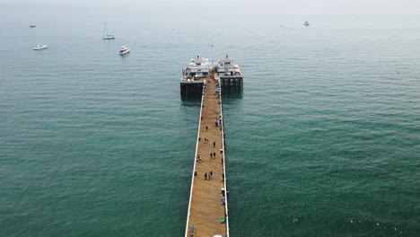 People-Walking-Along-the-Length-of-Malibu-Pier-with-Malibu-Farm-Cafe-at-the-End-of-the-Pier-and-Boats-in-the-Background-in-California-USA---Descending-Drone-Shot
