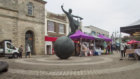 Drummer-Bronze-Sculpture-At-Lemon-Quay-With-Farmer's-Market-In-Background-In-Truro,-Cornwall,-UK