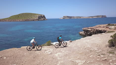 AERIAL---Two-people-ride-bicycles-next-to-the-ocean-in-Ibiza,-Spain,-wide-shot