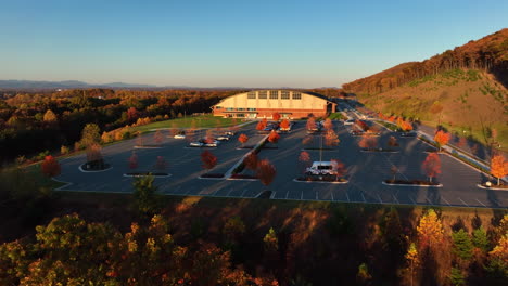Aerial-approach-of-large-sports-facility-building-and-parking-lot-on-side-of-mountain-during-autumn-fall-foliage