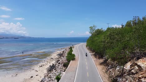 Aerial-drone-of-a-beautiful-coastal-road-and-the-ocean-with-a-scooter-motorbike-driving-around-a-blue-sky-sunny-day-on-tropical-island