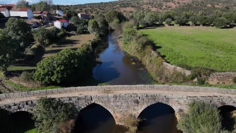 Drone-flies-over-Roman-bridge-next-to-a-small-river-town-in-Portugal