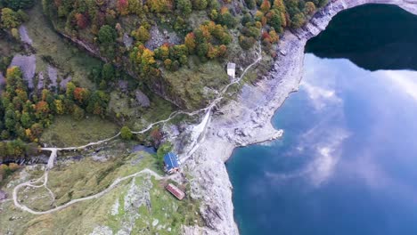 Lac-d'Oô-dam-stone-wall-and-keeper's-cabin-at-artificial-lake-in-the-French-Pyrenees-with-water-stream,-Aerial-top-view-orbit-around-shot