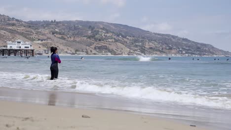 Mujer-Surfista-Parada-Al-Borde-De-La-Playa-Cuidando-A-Otros-Surfistas