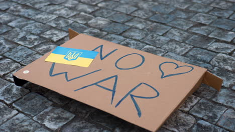 Antiwar-cardboard-sign-with-Ukrainian-flag-on-cobblestone-pavement