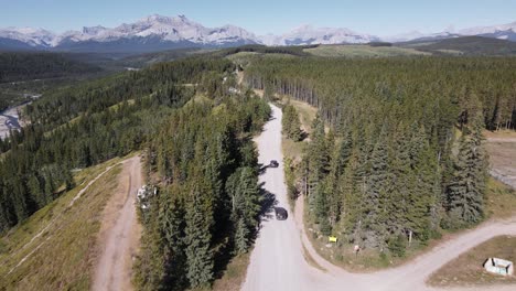 Two-cars-driving-along-a-dusty-logging-road-through-coniferous-forests-with-a-majestic-Rocky-Mountain-backdrop-near-Ghost-River