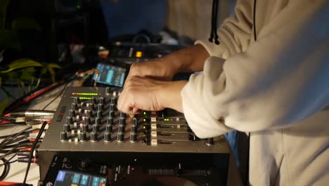 Roving-camera-films-hands-of-person-in-white-sweater-making-adjustments-to-DJ-mixer-and-console-during-live-DJ-performance