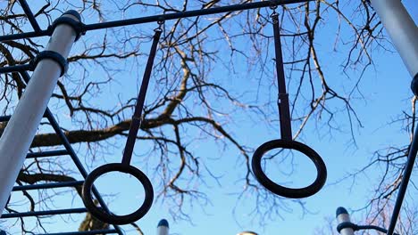 gymnastic-rings-move-in-the-wind-with-trees-and-blue-sky-on-the-background,-steady-close-up-shot