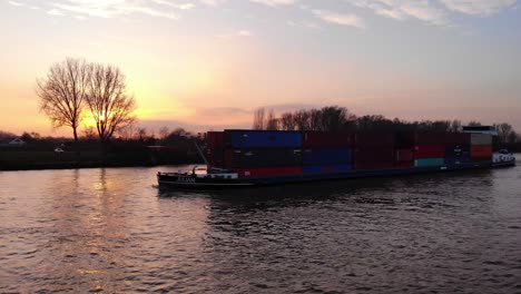 Luftaufnahme-Gestapelter-Container-Auf-Dem-Frachtschiff-Julia-Entlang-Der-Oude-Maas-Vor-Gelbem-Sonnenuntergangshimmel