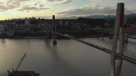 two-electric-public-transportation-trains-crossing-suspension-sky-bridge-spanning-over-river-connecting-light-rail-train-tracks-New-Westminster-Wide-angle-Aerial-ascending-panning-down-tracking