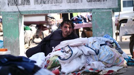 Dishevelled-Pakistani-Male-Lying-Under-Stone-Sign-Be-Stack-Of-Clothes-In-Front-Of-Him-In-Karachi