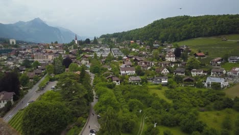 Aerial-dolly-out-of-picturesque-Spiez-green-valley-and-village-revealing-medieval-castle,-woodland-and-Swiss-Alps-in-background