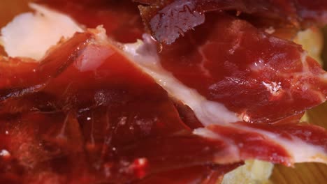 Macro-shot-of-oily-high-quality-spanish-serrano-ham-with-bread-rotating-on-wooden-board,-close-up-view-from-above