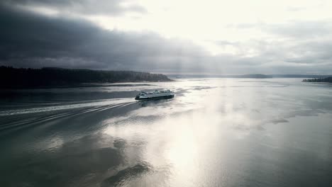 The-brilliant-morning-sun-streams-through-dark-gloomy-clouds-as-a-ferry-passes,-Aerial-orbit