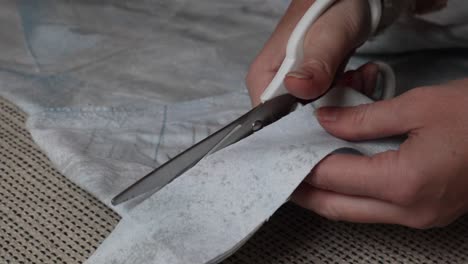 Closeup-of-young-woman-hands-carefully-cutting-light-blue-fabric-cloth-material-with-white-scissors