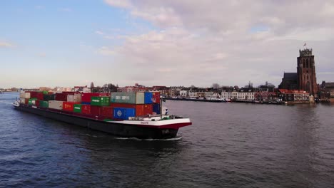 Aerial-Starboard-Side-View-Of-Salute-Cargo-Ship-Going-Past-On-Oude-Maas-With-Our-Lady-of-Dordrecht-In-Background