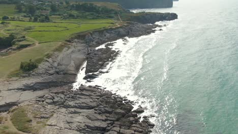 A-drone-rises-slowly,-over-the-sea-as-the-waves-crash-relentlessly-against-jagged-grey-rocky-shoreline,-revealing-endless-lush-fields-of-green
