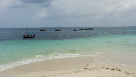 Boats-anchored-just-offshore-white-sand-beach-in-Zanzibar,-one-sailing