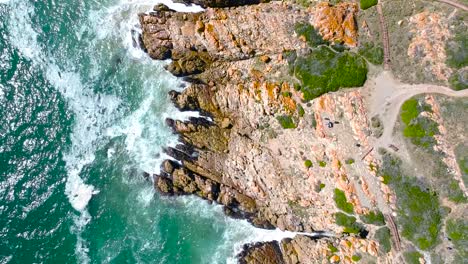 Top-Down-View-of-rocky-coastline-with-turqoise-waves-crashing-into-the-rocks-on-a-sunny-day