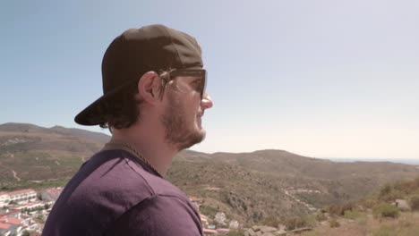 Man-with-baseball-hat-and-sunglasses-walking-around-hills-on-sunny-summer-day