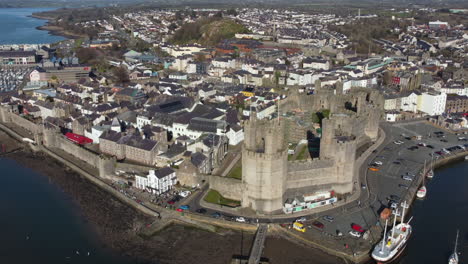 An-aerial-view-of-Caernarfon-Castle-on-a-sunny-day,-flying-left-to-right-around-the-castle-while-zooming-in,-Gwynedd,-North-Wales,-UK