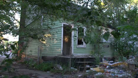 Dilapidated-wooden-house-with-broken-windows-and-no-door-and-huge-amounts-of-garbage-in-the-yard-among-the-green-trees-in-Detroit