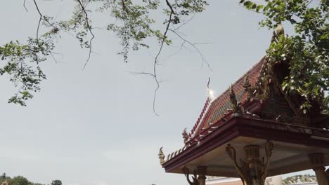 Static-shot-of-a-thai-temple-roof-shining-on-sun-and-tree-branches-swaying-on-wind