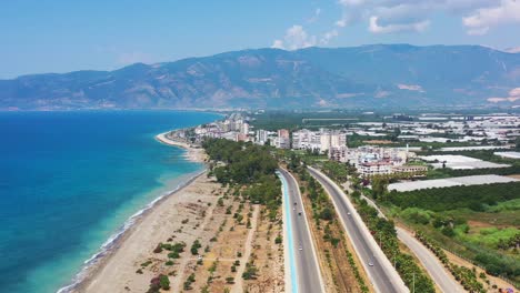 Wide-aerial-view-of-an-empty-highway-road-leading-a-coastal-beach-town-with-tropical-blue-Mediterranean-Sea-and-large-mountains-in-the-distance