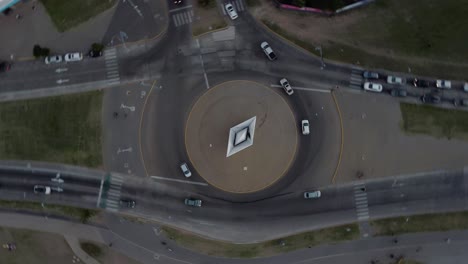 Aerial-drone-top-rotating-shot-over-a-road-crossing-in-Rosario,-Argentina-during-evening-time-with-cars-from-all-sides-passing-by-on-the-sides-of-a-white-boat-statue