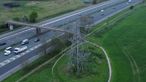 Vehicles-on-M62-motorway-passing-pylon-tower-on-countryside-farmland-fields-aerial-view-following-traffic-right