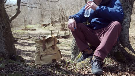 Older-man-with-rucksack-sits-by-tree-in-forest-drinks-water