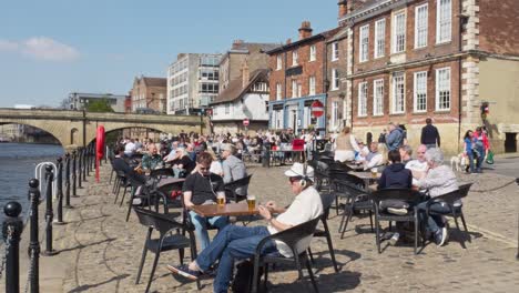 People-relaxing-and-enjoying-drinks-at-a-riverside-bar-in-the-ancient-city-of-York-UK-England-during-a-bright-sunny-day