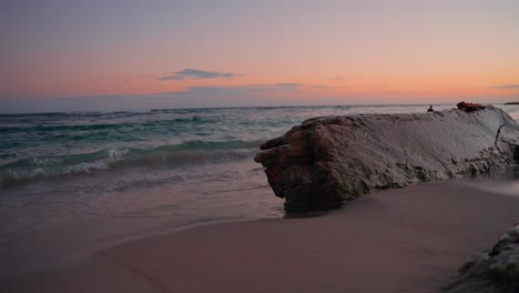 Statische-Ansicht-Des-Von-Meereswellen-Nassen-Felsens-Am-Strand-Bei-Sonnenuntergang-Mit-Der-Silhouette-Einer-Person-Im-Hintergrund