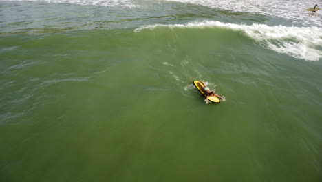 An-aerial-shot-of-a-surfing-woman-lying-on-a-yellow-surfboard-and-swimming-in-direction-of-a-wave