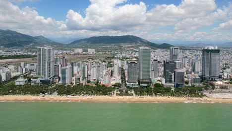 aerial-drone-flying-parallel-to-the-white-sand-beach-coastline-full-of-palm-trees-and-white-beach-umbrellas-in-Nha-Trang-Vietam-on-a-sunny-day-with-tall-residential-buildings-along-a-coastal-road