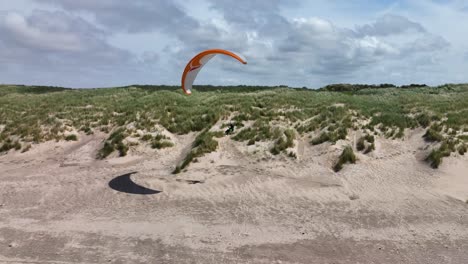 Aerial-shot-of-a-paraglider-riding-the-lift-band-of-a-green-grass-covered-dune-with-white-sand-on-a-sunny-day-by-the-sea-at-Ouddorp,-Brouwersdam,-Netherlands