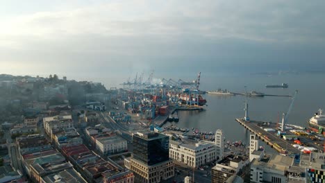 Aerial-jib-up-over-buildings-near-cranes,-containers-and-ships-docked-in-Valparaiso-Sea-Port-on-a-cloudy-day,-Chile