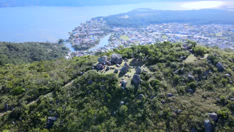 Aerial-drone-scene-of-mountain-with-rocks-on-top-with-city-between-ocean-and-river-in-background