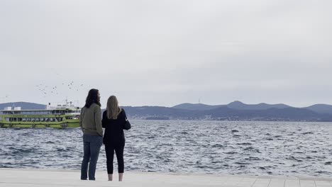 Ein-Verliebtes-Paar,-Das-Vorbeifahrende-Boote-Auf-Einer-Wunderschönen-Promenade-In-Zadar,-Kroatien,-Beobachtet