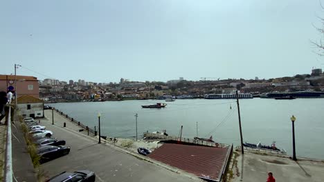 A-Vessel-Cruising-The-Placid-Waters-In-Porto,-Portugal-With-View-Of-Parked-Cars-And-People-On-The-Forefront-And-Coastal-City-Structures-In-The-Background