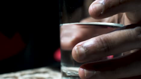 A-close-up-shot-of-the-hand-of-an-ethnic-man-holding-a-glass-filled-with-a-clear-liquid,-the-man-fidgeting-by-rotating-the-glass-on-the-table-with-his-fingers