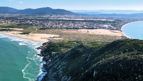 Aerial-drone-scene-of-urbanized-paradise-beach-with-sand-dunes,-mountains-and-sea-beach-of-santinho-Florianópolis-mountain-top-and-city-on-the-horizon