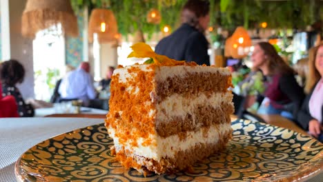 Delicious-carrot-cake-with-a-yellow-flower-on-a-plate-in-a-pretty-restaurant,-sweet-dessert,-unrecognizable-blurry-people-in-the-background,-4K-static-shot