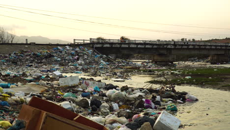 Garbage-piled-up-on-river-banks,-motorbikes-pass-over-bridge,-Vietnam