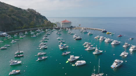 Flying-Over-Beautiful-Catalina-Island,-Boats-Sitting-on-Clear-Turquoise-Waters-of-Avalon-Harbor